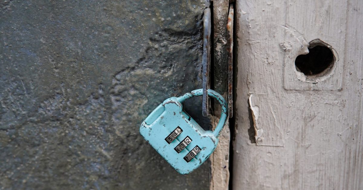 A small, turquoise-colored combination padlock hangs from a rusty metal latch on a weathered door
