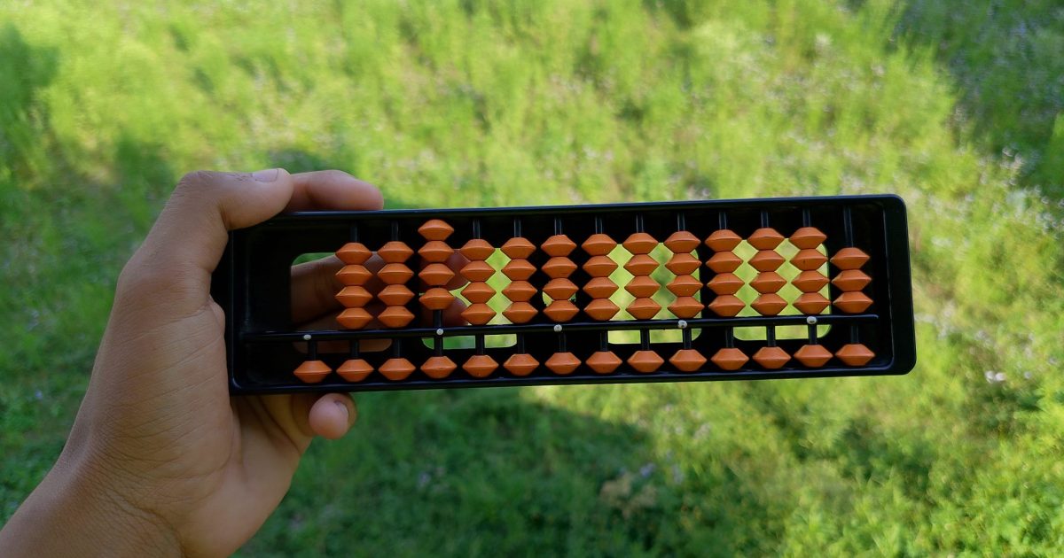 A hand holds a black abacus with orange beads against a background of lush green grass