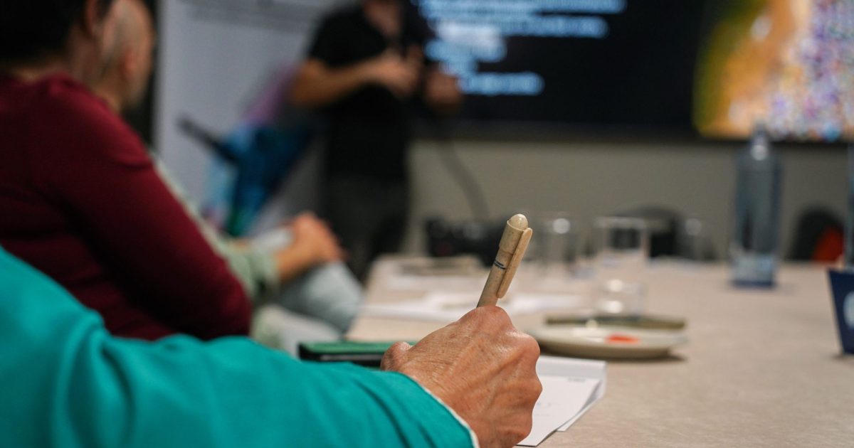 A person holding a pen is taking notes at a table during a presentation